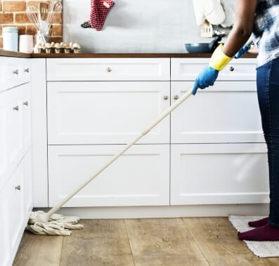 Person in blue gloves mops wooden kitchen floor with string mop, white cabinets.
