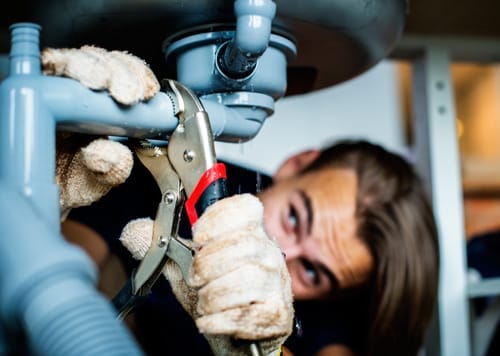 Person in gloves uses wrench to fix light blue plumbing pipes under a sink.