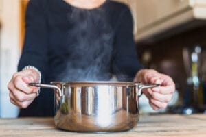 Hands hold a steaming stainless steel pot on a wooden kitchen counter.