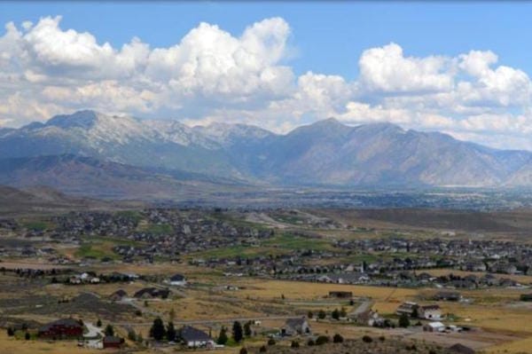 Suburban sprawl in a valley before vast, rugged mountains under a blue, cloudy sky.